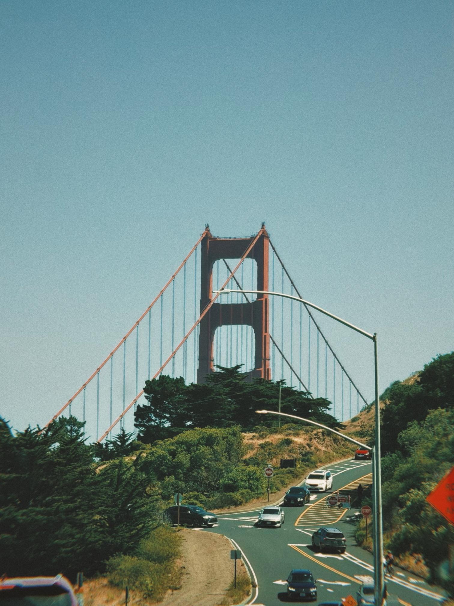 A view of the Golden Gate Bridge in the background with vehicles on a winding road in the foreground, surrounded by greenery and blue sky.