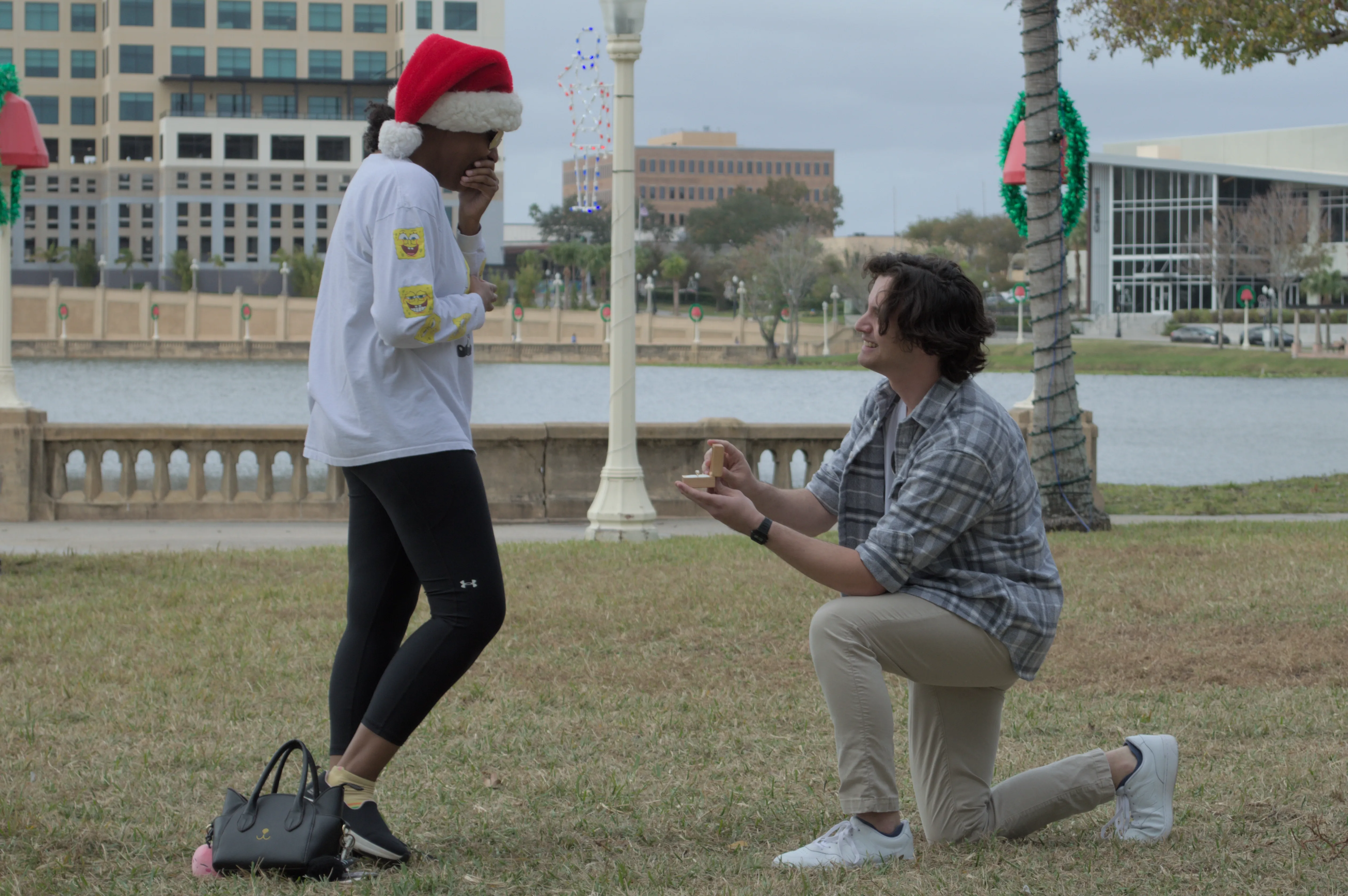 Josh on the right kneeling down presenting the ring to Amaya, who is covering her mouth in shock and delight.