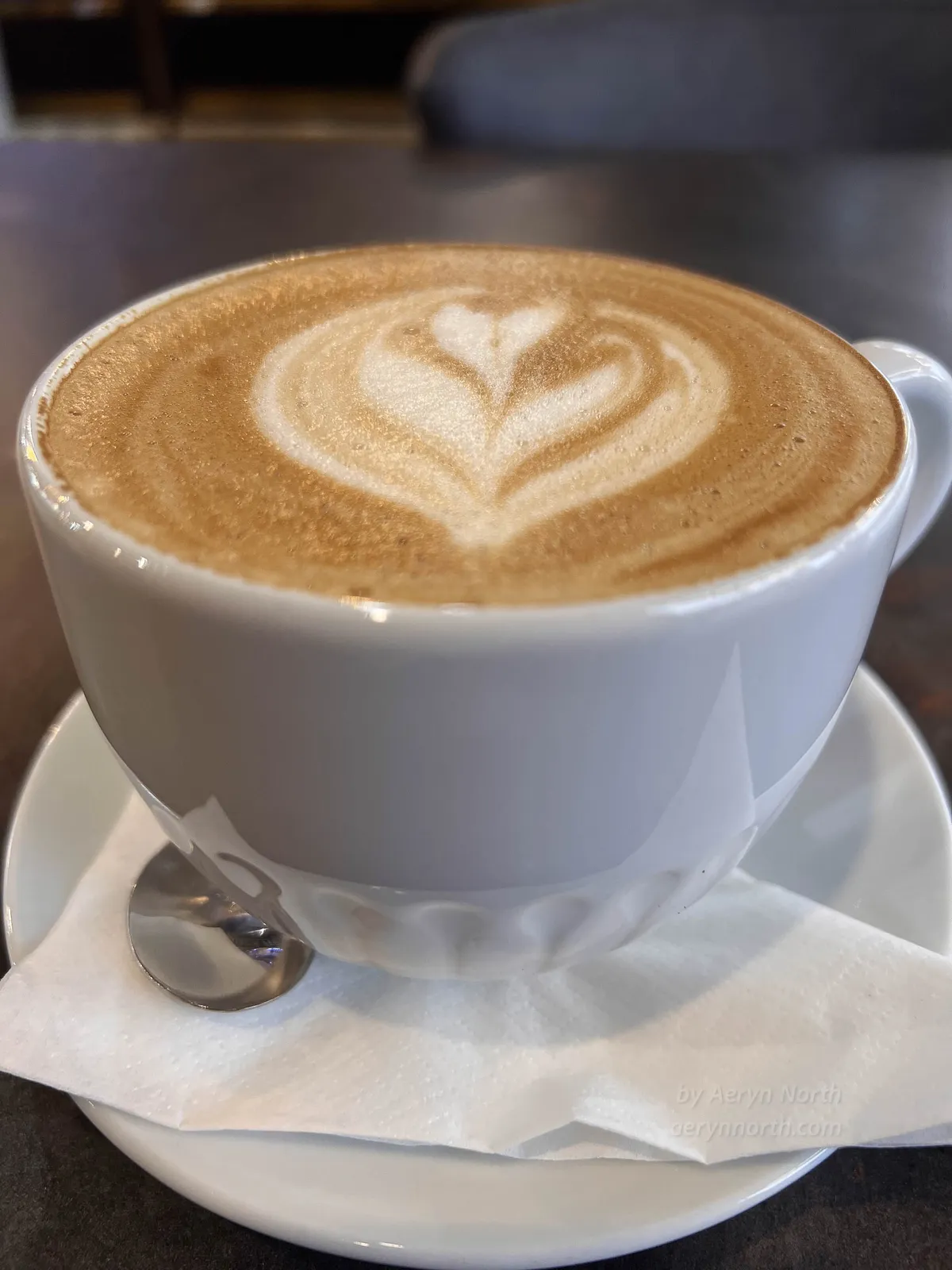 On a brown table a latte with latte art on the top sits in a saucer with a napkin.