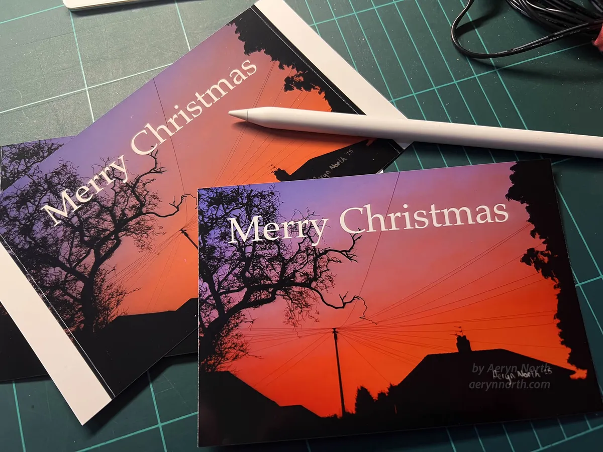 Postcard-sized photographs lie on a green cutting mat next to a white Apple Pencil. The photographs feature a scene of a cloudy sky backlit by pink, orange, and lilac hues from the sunrise. In the forground, trees, houses, and a telephone pole with radiating cables are silouetted against the sky.
