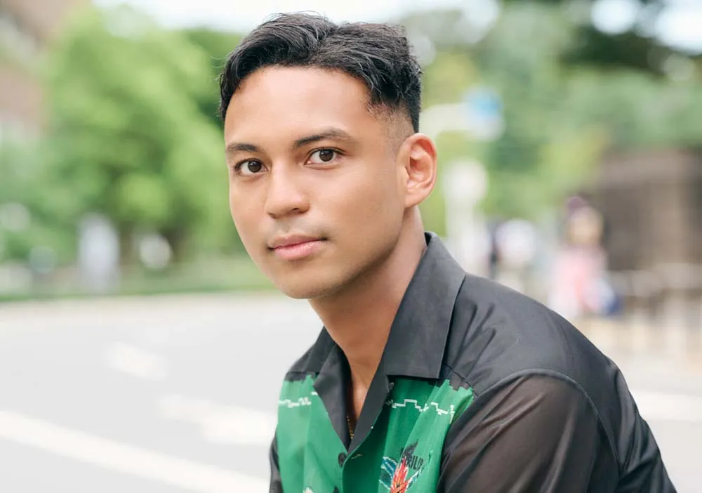 A picture of author Jose Ando. He is a man of Japanese and African descent, with dark brown skin, very dark brown eyes, and black hair cut short on the top and buzzed tight on the sides and back. He is wearing a black and green collared shirt and turned from a 3/4 perspective to look at the camera.