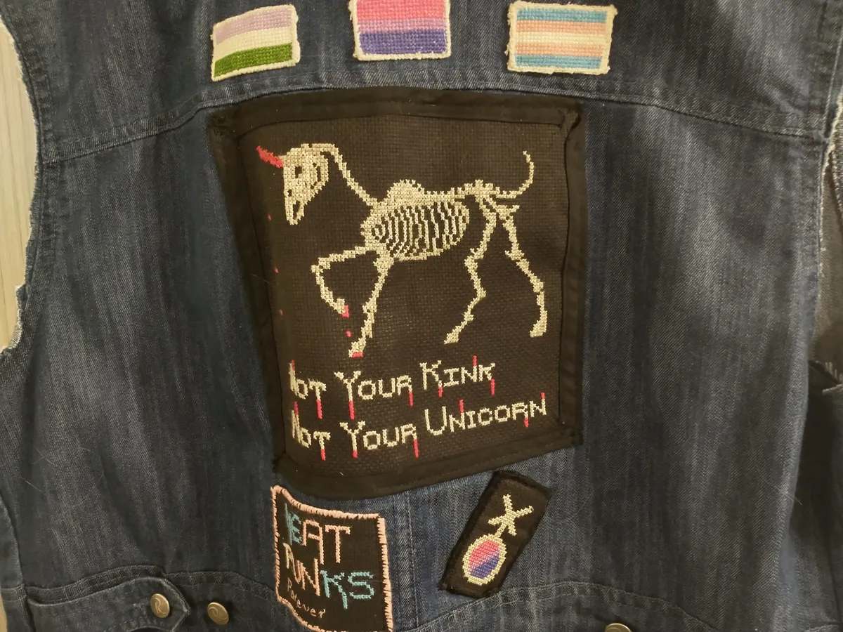 Photograph of denim protest vest with multiple homemade patches. The central patch shows a skeletal unicorn with a bloody horn with the text "Not your kink, not your unicorn."
