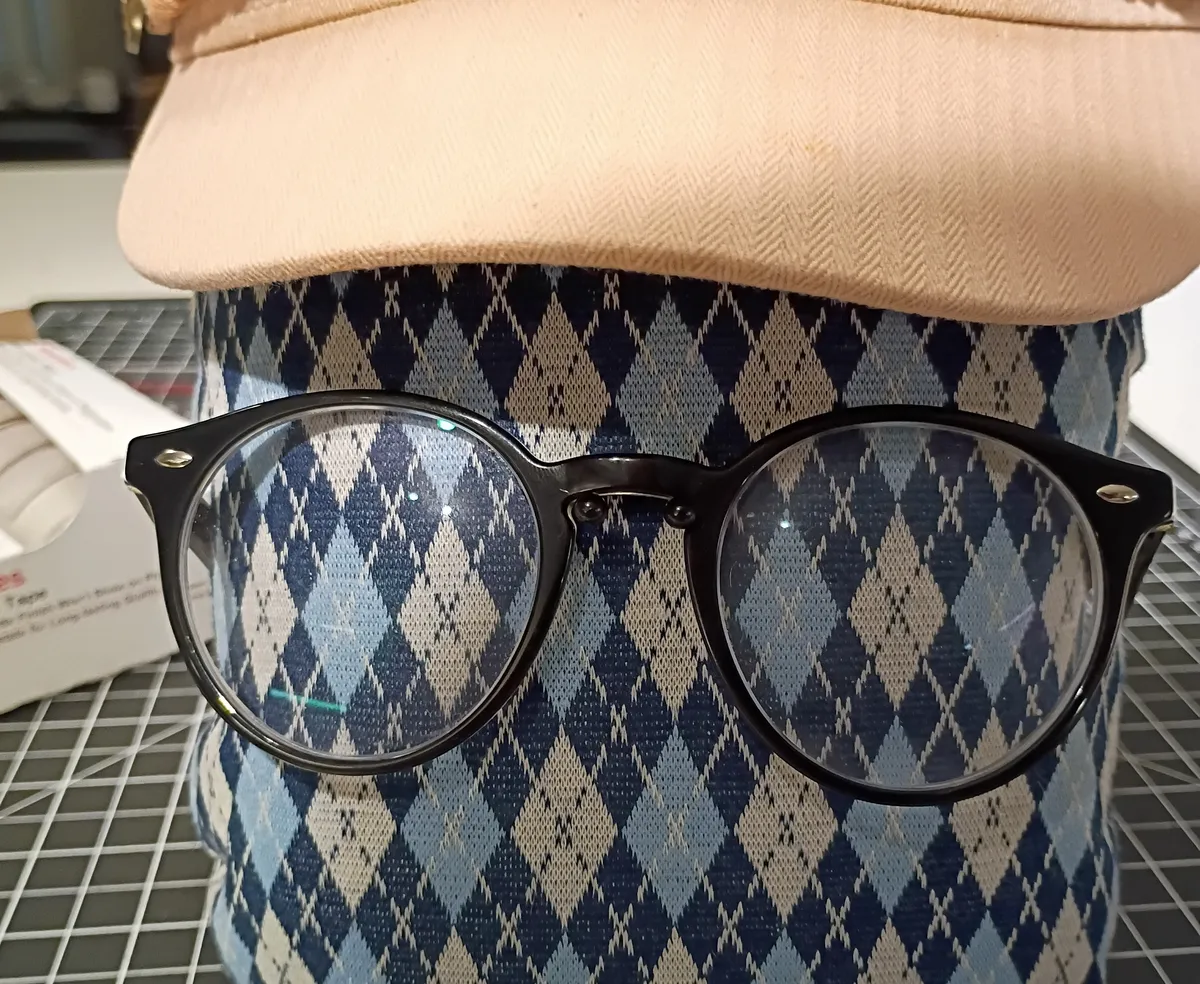mixed media sculptural selfie consisting of a tailor's ham with a pair of black glasses pinned to it and a pink hat with a brim perched on top
