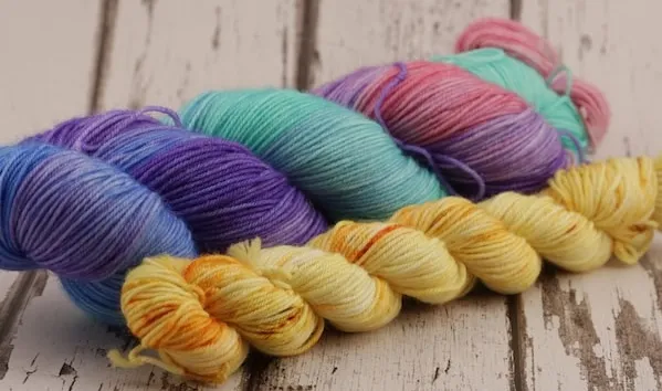 Two skeins of yarn on a weathered wooden surface. The one in the back is a large skein of sock wool in blue, teal, purple, and pink colours. The one in the front is a much smaller skein, mostly in gold.