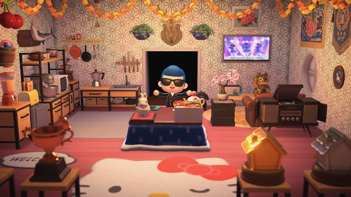Screenshot from Animal Crossing: new Horizons. Slothra is wearing a black leather jacket, a pair of black sunglasses, and has blue hair put up into a pompadour. They are standing behind a kotasu that has a lot of clutter on top. To the right of them is a mid-century stereo console, a box of vinyl records, a gold lucky cat, a cherry tree bonsai, and a television on the wall. To the left of them is a kitchenette with a sink, microwave, moka pot, mixer, toaster, and blender. There is a lot of art and banners on the walls, and a large Hello Kitty rug right in the entry way.