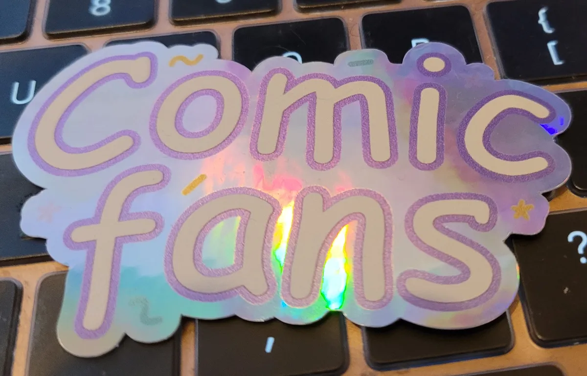A holographic sticker that says "Comic Fans" in the Comic Sans typeface