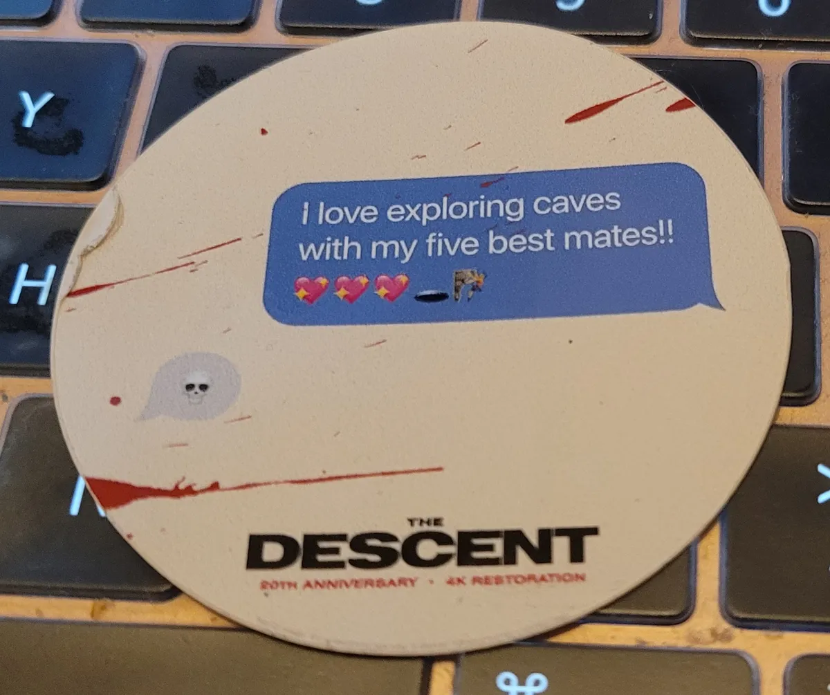 A circular sticker which says at the bottom "The Descent", "20th Anniversary", and "4k Restoration". In the main part of the sticker, there is a text bubble that says "I love exploring caves with my five best mates!!" and three sparkly heart emojis, a hole emoji, and a rock climber emoji. The response to that text is another text bubble with just a skull emoji in it. There is also blood spatter across the sticker.