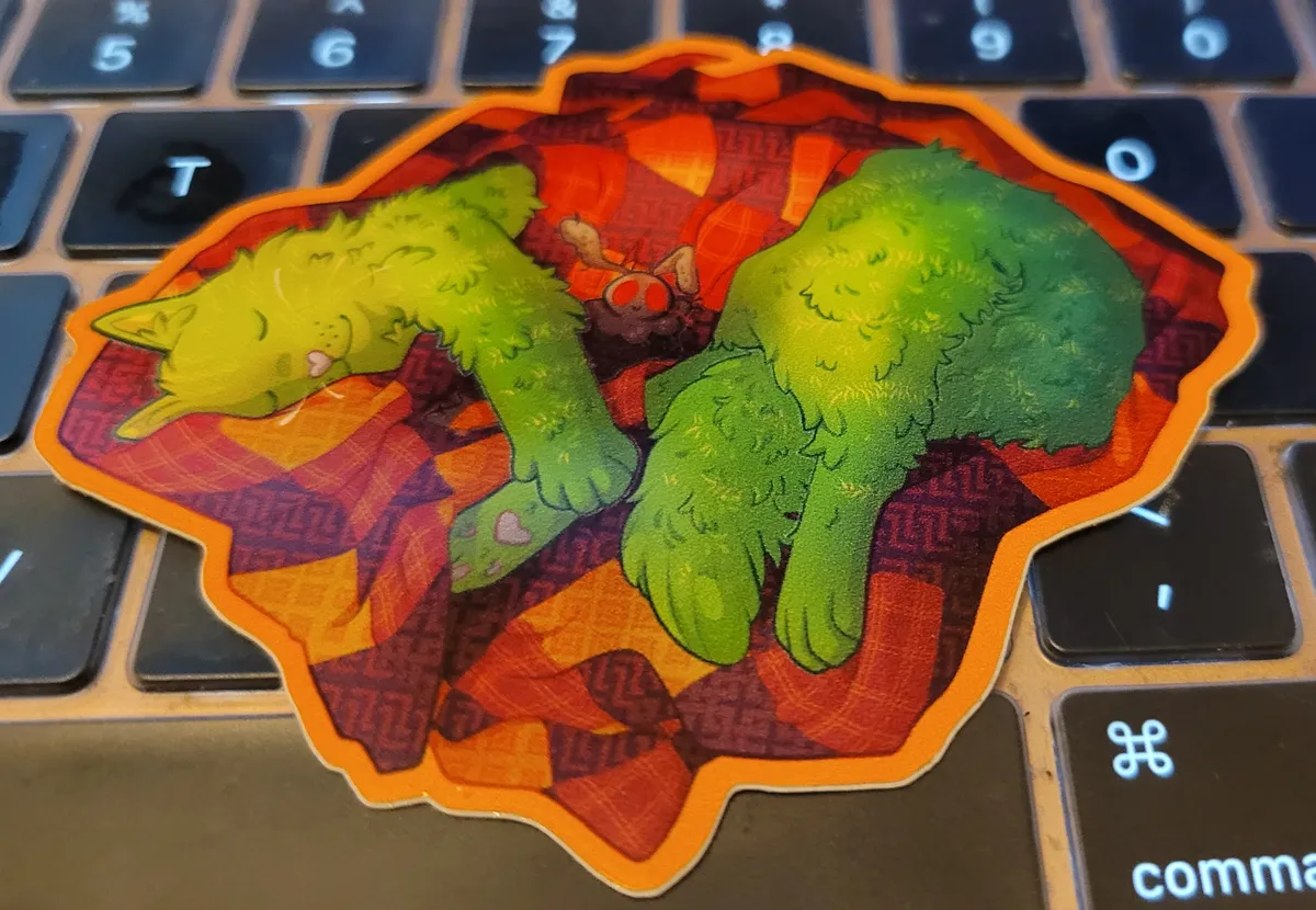 A cactus cat, green and prickly, is dozing comfortably under a patchwork blanket, with a small Mothman plushie next to her.