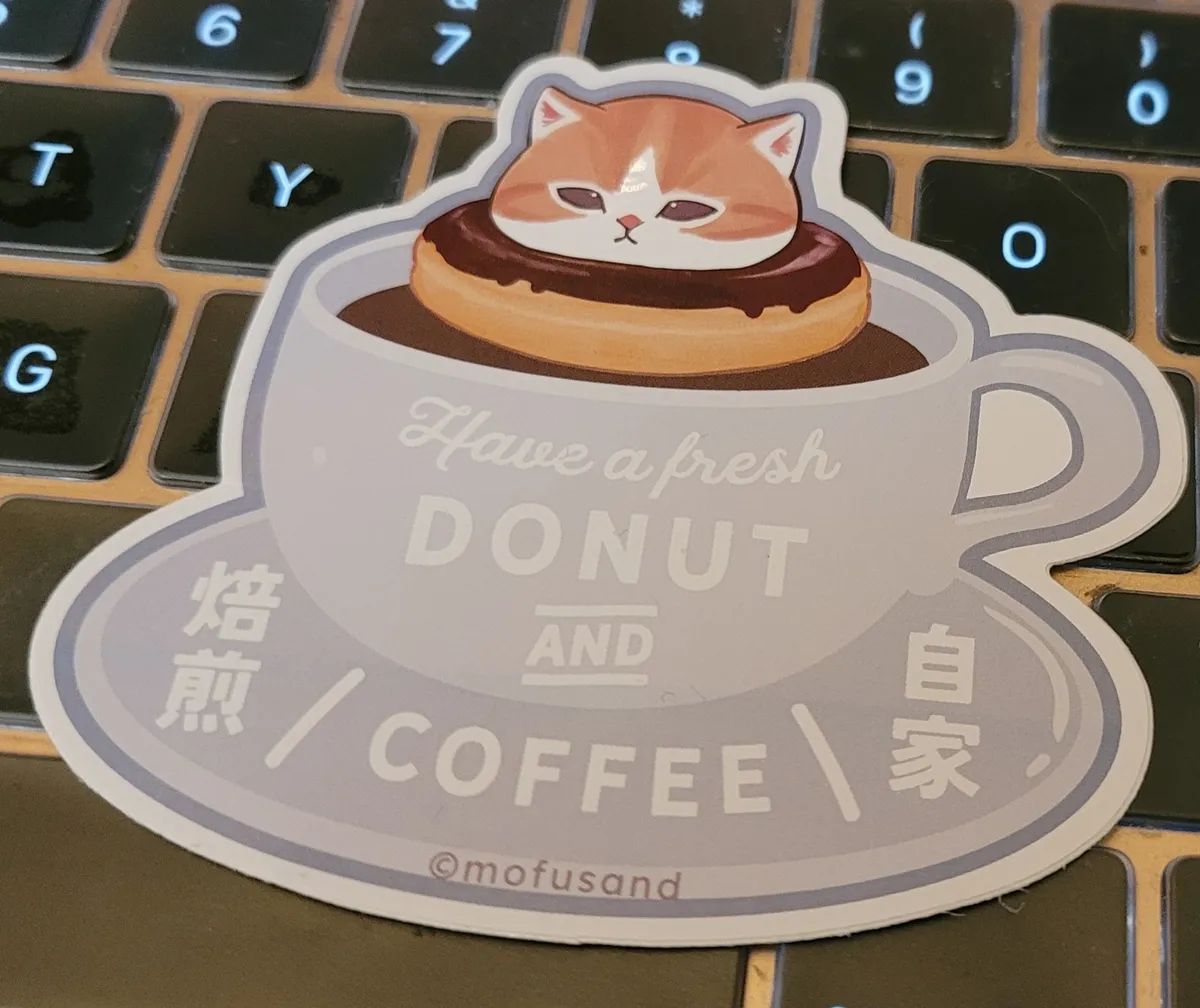 An illustration of an orange and white cat, with a chocolate-glazed donut around its neck, floats in a cup of coffee. The coffee cup and saucer say "Have a fresh donut and coffee" as well as "Roasting House" in Japanese Kanji. Underneath all of that is the copyright symbol and "Mofusand", who was the designer.