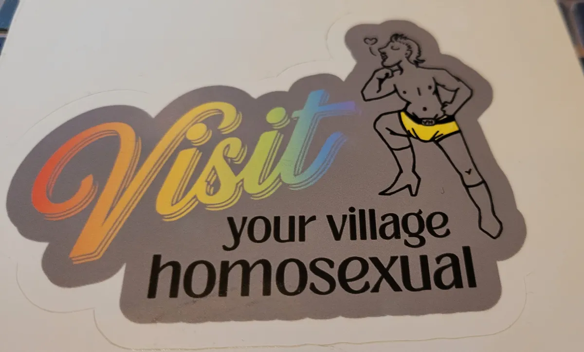 On a grey background is the phrase "Visit your village homosexual", with "visit" in a rainbow gradient. On the right, with one foot resting above "village" is a man wearing heeled boots and a bright yellow pair of tiny shorts (and that's it). He is pursing his lips and there is a little heart word balloon.
