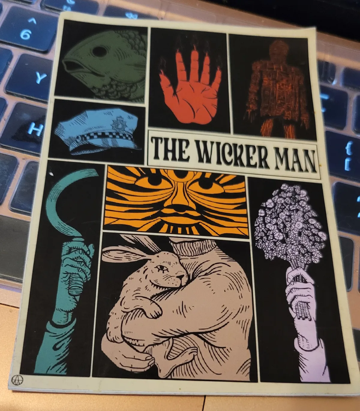 A sticker with a selection of iconic images from the original The Wicker Man film, including the Salmon of Knowledge, the Hand of Glory, the Green Man mask, a hare, a police officer's hat, and a sickle held aloft. There is also the titular Wicker Man.