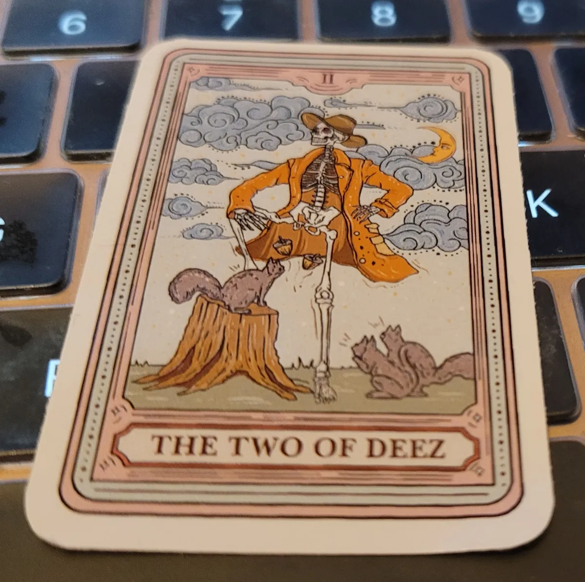 An illustration of a Tarot card, where a skeleton wearing a large flappy coat and a flappy hat is standing, his left leg up on a tree stump, as two acorns fall from his pelvic bones towards a waiting squirrel, who is also perched on that tree stump. Two other squirrels are looking at him in amazement from the right, and below this picture is the phrase The Two of Deez.