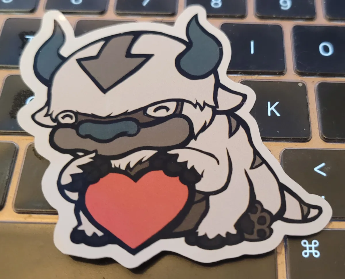 Appa, the sky bison from Avatar: The Last Airbender, is sitting on his back two paws, using his middle two paws to support him, and holding a heart between his front two paws.