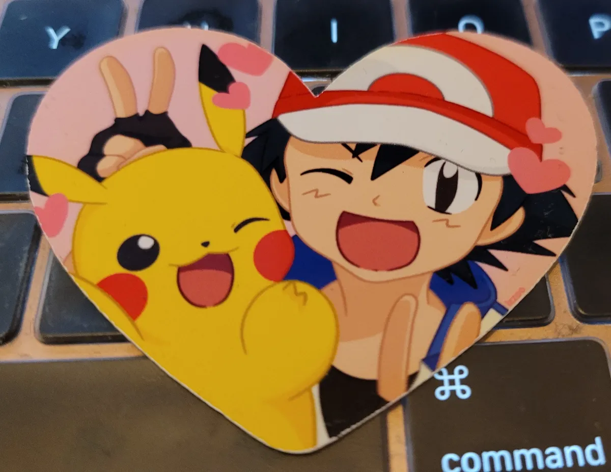 A heart-shaped sticker with Ash and Pikachu winking and giving the V-for-victory fingers