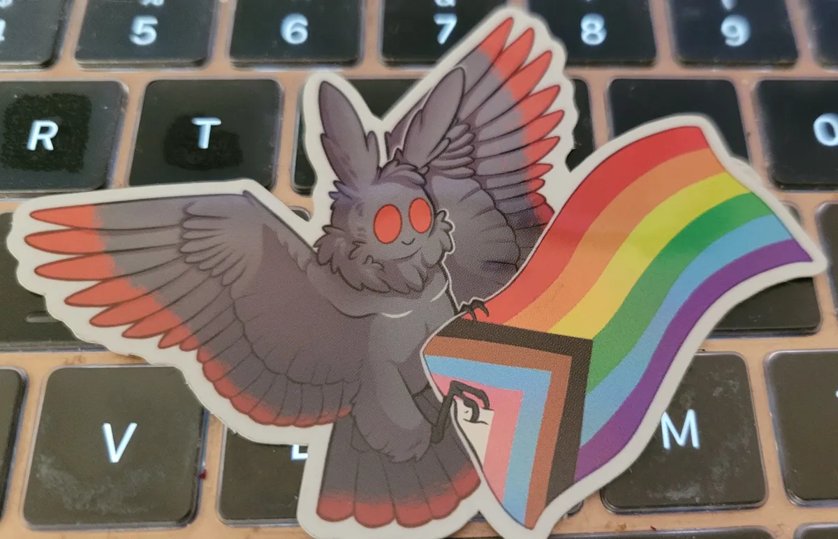 A shiny metallic sticker of Mothman holding an inclusive Pride flag in his tiny little claws