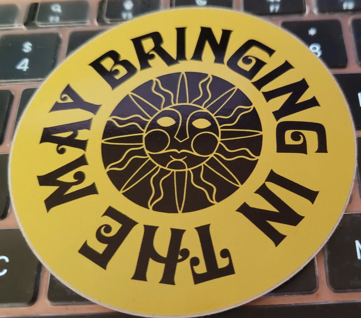 A bright yellow circular sticker, with an illustration of the sun in the middle and around it the words "Bringing In The May"