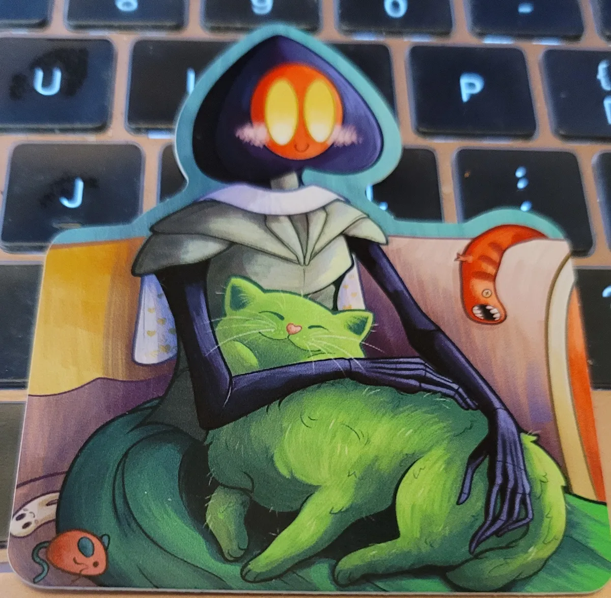 An illustration of the Flatwoods Monster holding a Cactus Cat while sitting on a couch. A Mongolian Death Worm plushie is draped over the back, and a small toy mouse and toy Fresno Nightcrawler are sitting next to her.