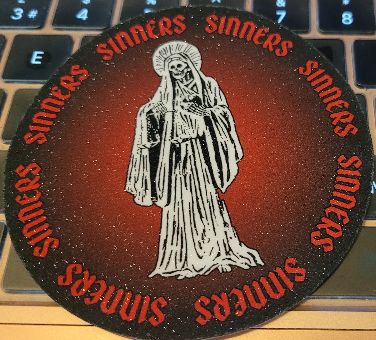 A sparkly circular sticker with a black background. There's an illustration of Santa Muerte in the middle (a female saint with a skull for a face), and all around the edge it says 