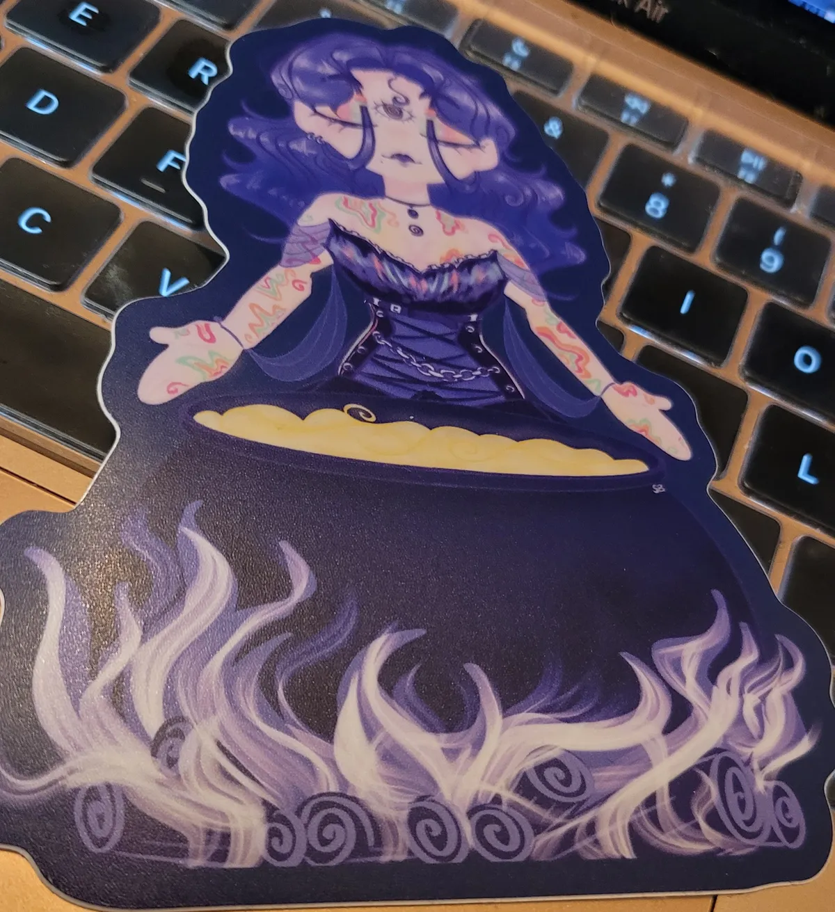 An illustration of a witch with purple hair, tattoos, and wearing a purple corset top, who is standing behind a bubbling cauldron, heated by a wood fire underneath. Her eyes are closed and her arms are outstretched in supplication.