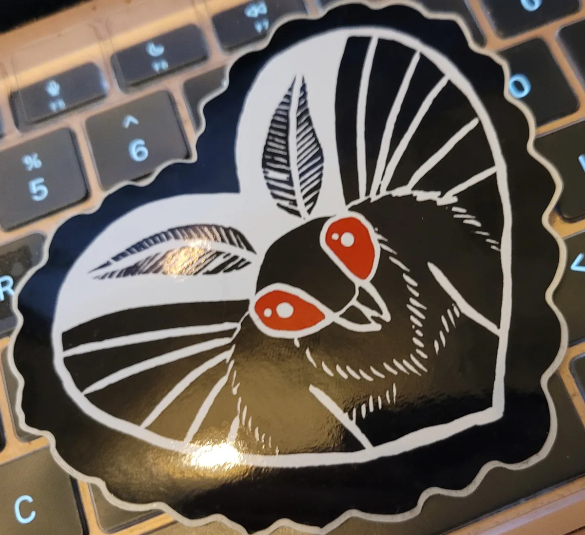 A large heart-shaped sticker with a Lino-cut illustration of Mothman inside a lacy heart.