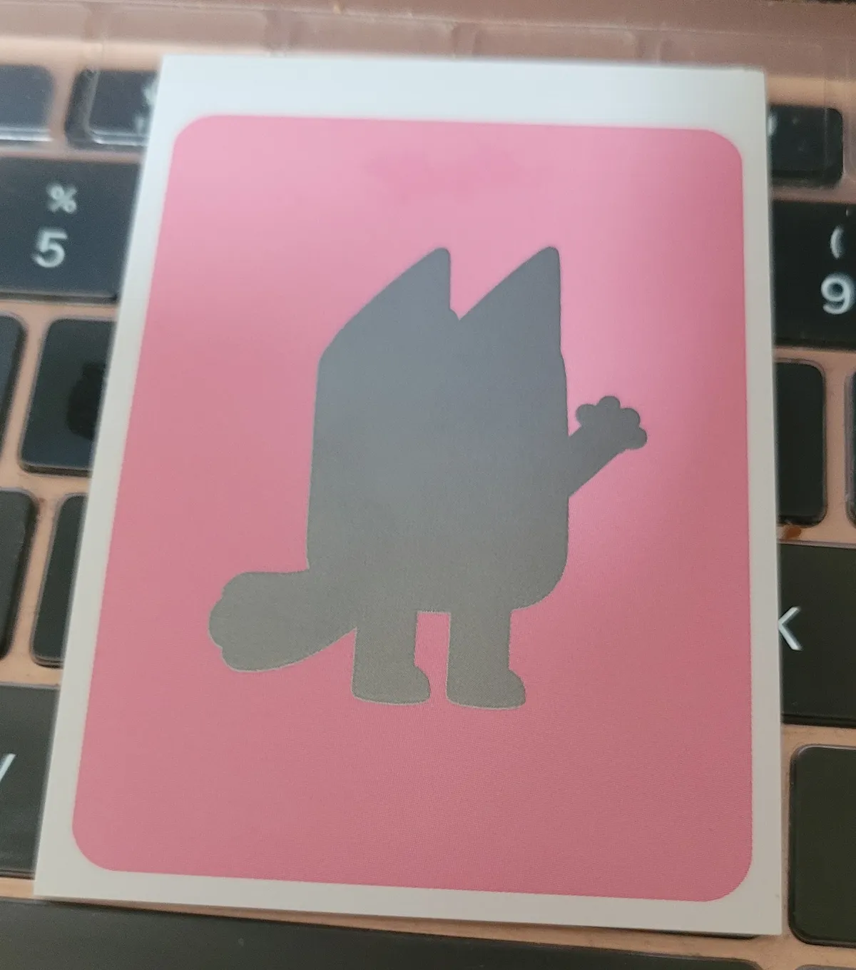 A silver silhouette of a dog from Bluey on a pink background.