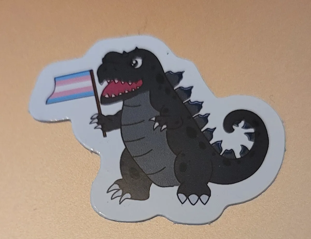 A drawing of Godzilla smiling widely and holding up a trans flag