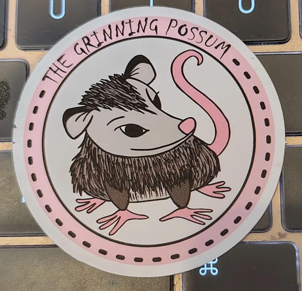 An circle illustration of a very cute possum with text around it that says The Grinning Possum. It could be a pub sign with how cute it is.