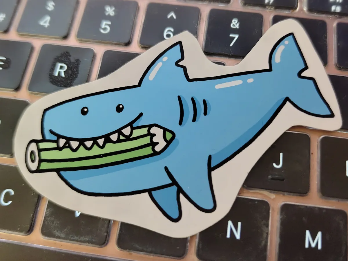 A shark is holding a green coloured pencil in its mouth and grinning at the same time.
