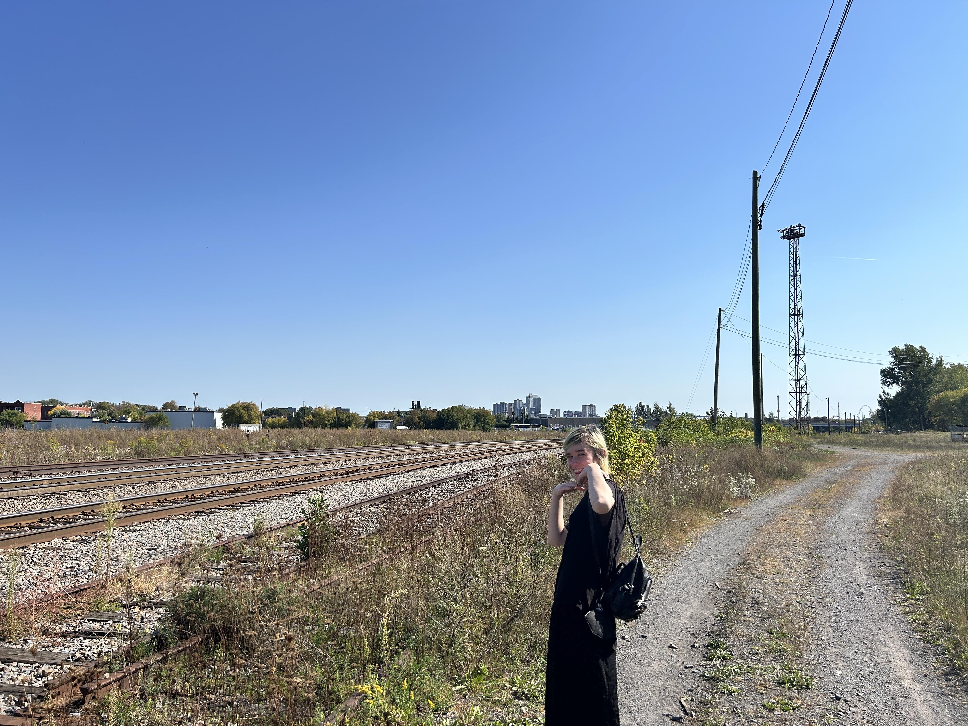 Noé standing out in a field next to some train tracks, posing with her hands under her chin framing her head. She's also very small and everything around her is very large.