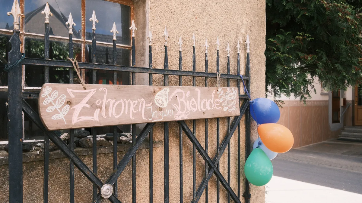 A handmade wooden sign reading “Zitronen Ingwer Biolade” hangs on a black wrought iron fence outside a rustic building. Several colorful balloons are tied to the fence beside the sign. The scene has a warm, village fair atmosphere, with weathered walls, a window reflecting nearby buildings, and greenery in the background.