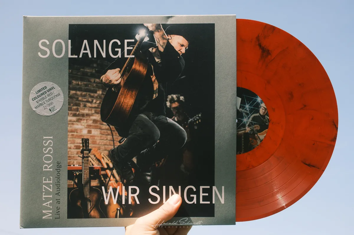 Matze Rossi — Solange wir singen, ist es für immer. marble red vinyl half out of the sleeve, held against a blue sky.