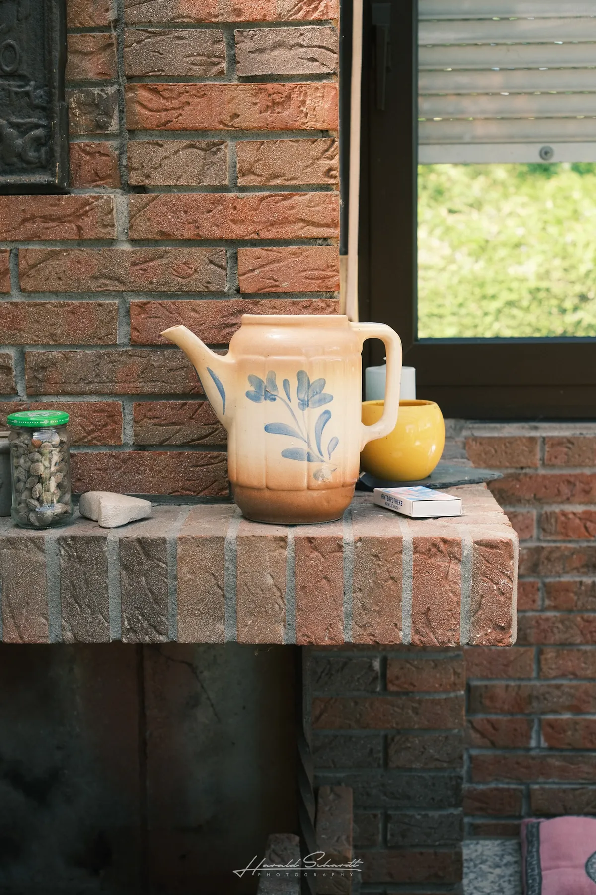 An old ceramic jug with a blue floral pattern on a brick fireplace ledge, a yellow pot and matchbox beside it.