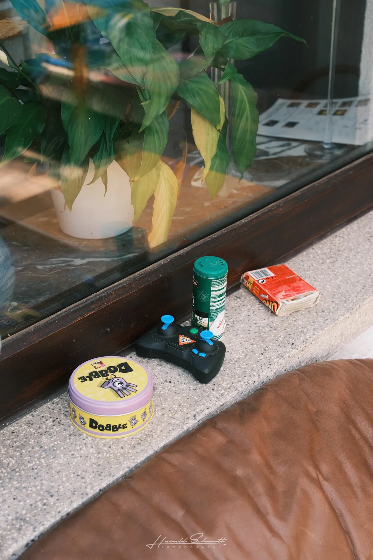 A window sill with a Dobble tin, a toy controller, a bug spray can, and a battered UNO box, houseplant reflected in the glass behind.