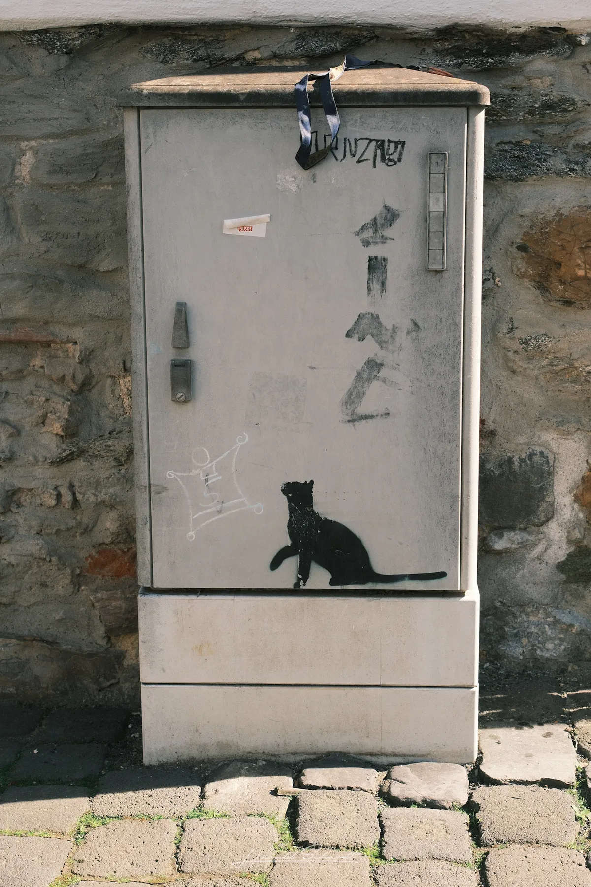 A grey utility box with a black stencilled cat and graffiti tags, set against a stone wall on cobblestones.