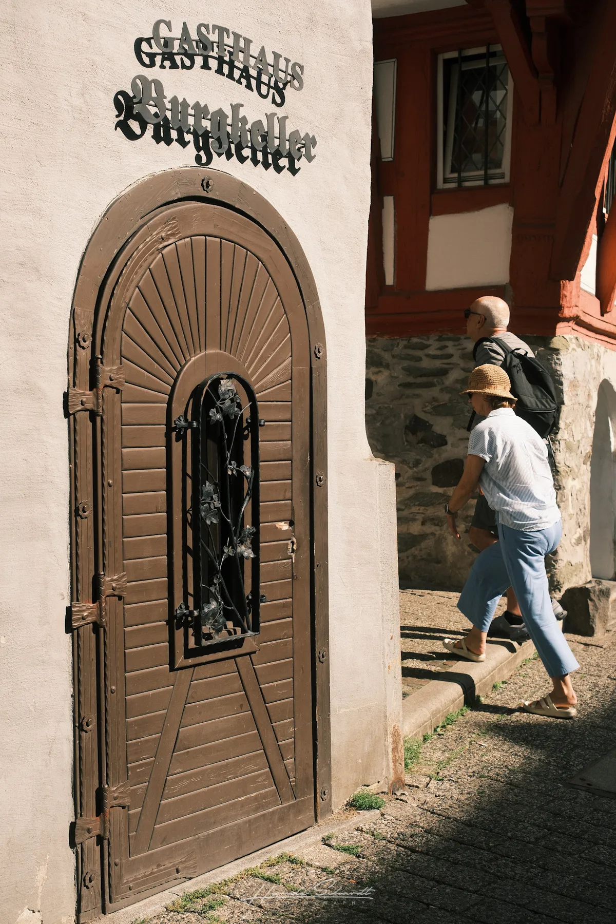 The arched wooden door of Gasthaus Burgkeller in Limburg old town, two tourists walking past in bright sunlight.