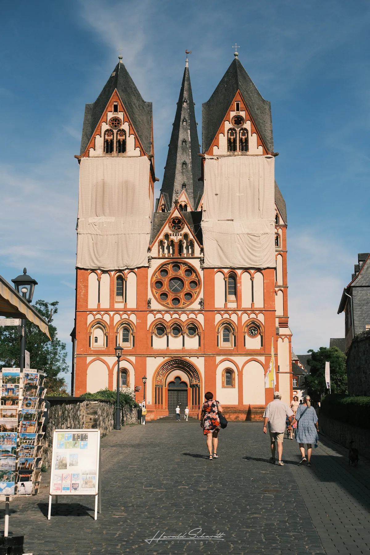 The facade of Limburg Cathedral with two towers wrapped in scaffolding cloth, tourists walking toward the entrance.