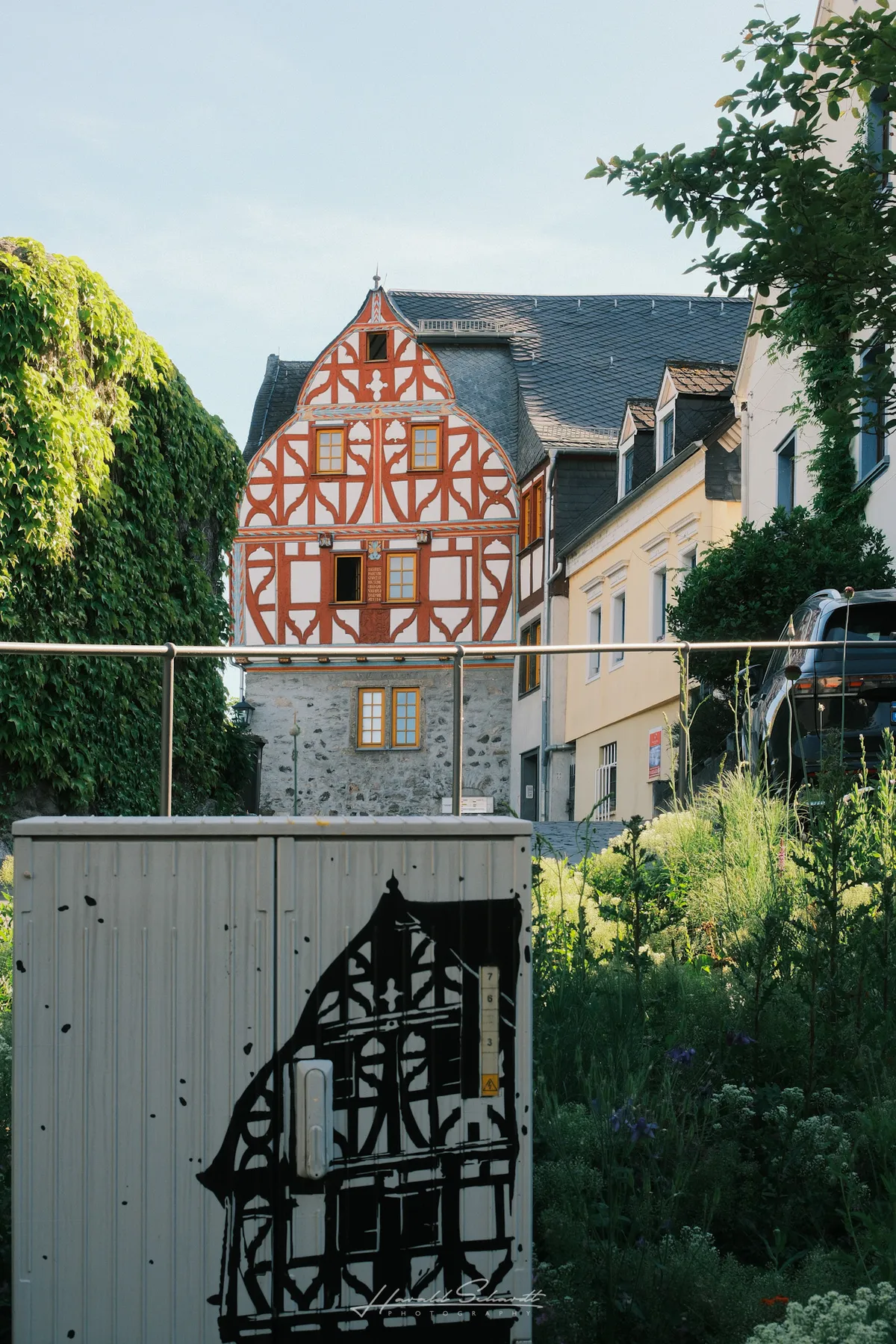 A ornate half-timbered house in Limburg, with a utility box in the foreground painted with a black stencil of the same building.
