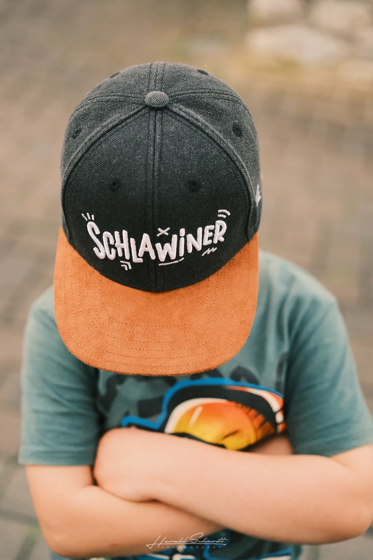A child with arms crossed, face hidden under a snapback cap reading Schlawiner, grey gravel background.