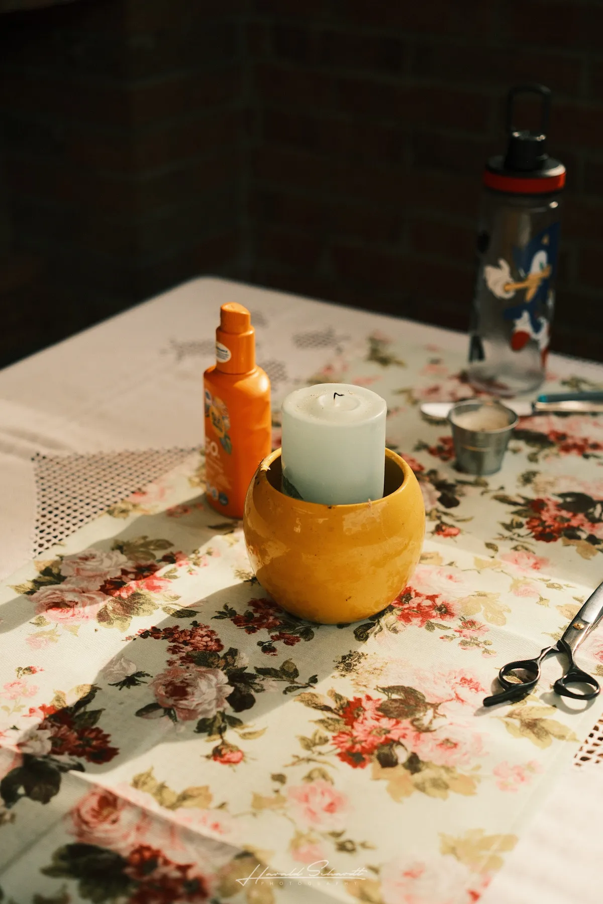 A yellow ceramic pot with a white candle on a floral tablecloth, sunscreen bottle and Sonic water bottle behind it.