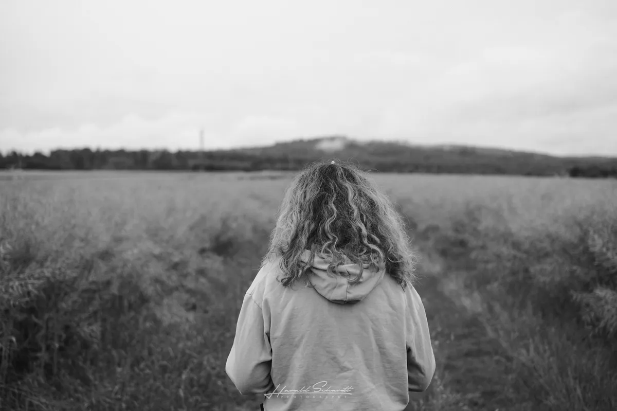 A woman standing on a path between fields, viewed from behind beneath a wide cloudy sky.