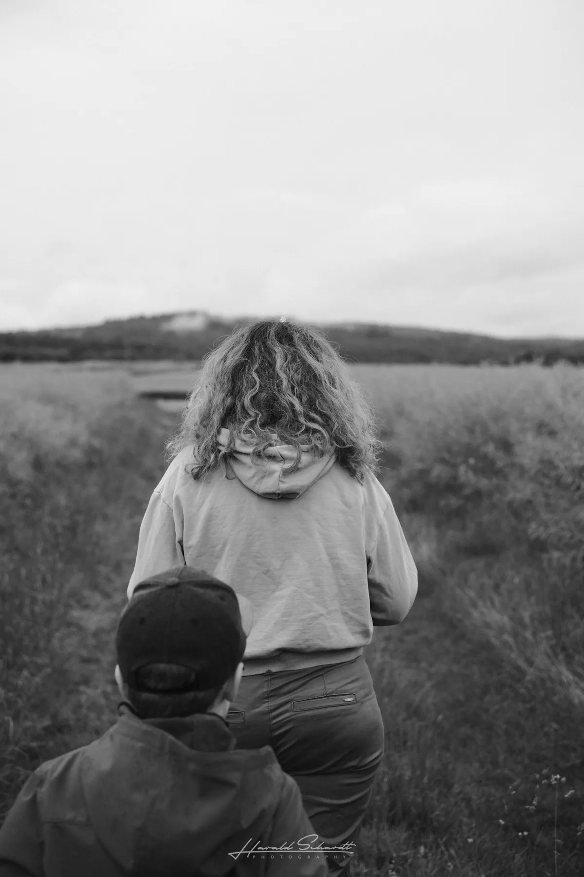 A woman and a child standing on a narrow path through a field, seen from behind under an overcast sky.