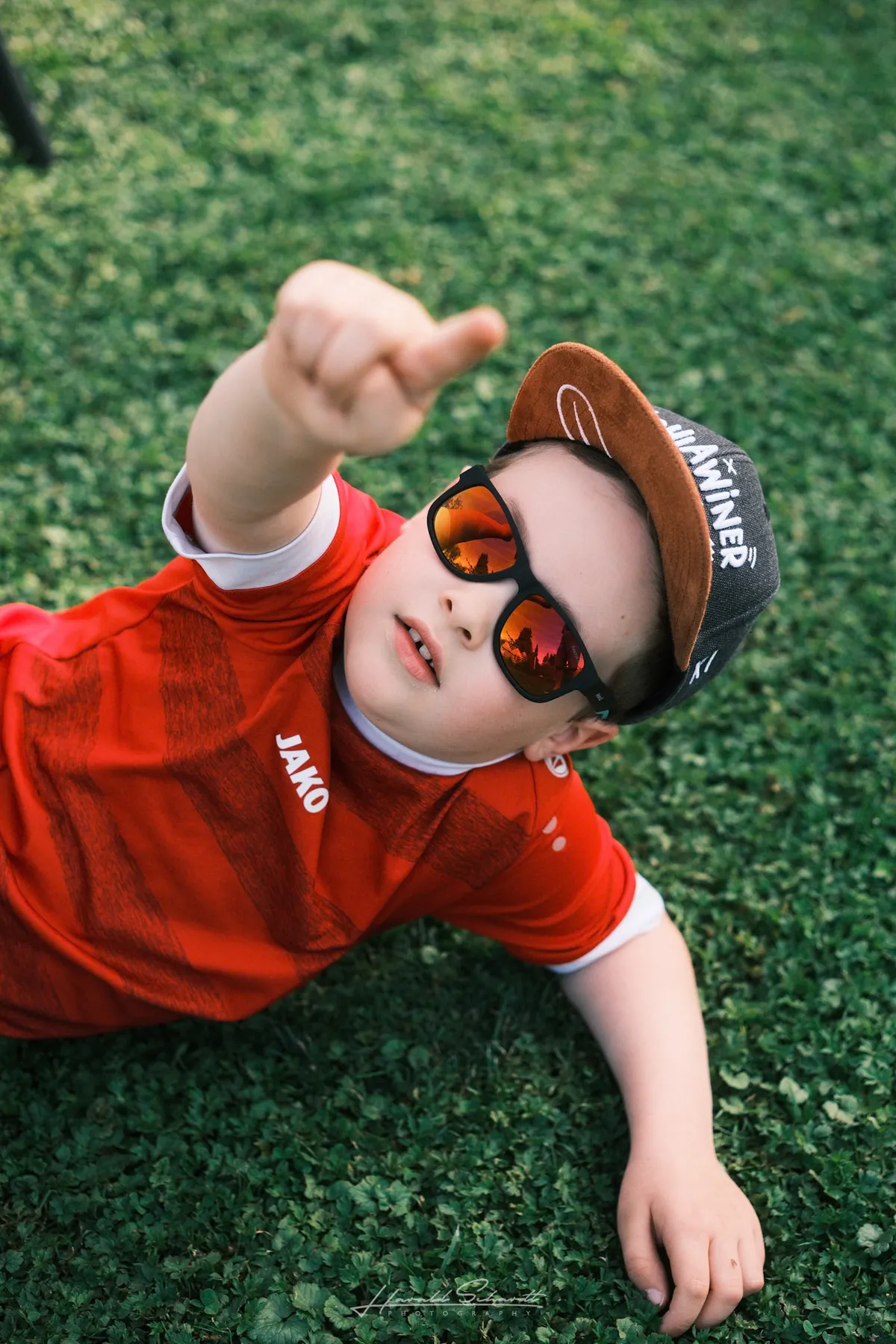 A young boy wearing sunglasses and a red jersey lies on the grass, pointing toward the sky.