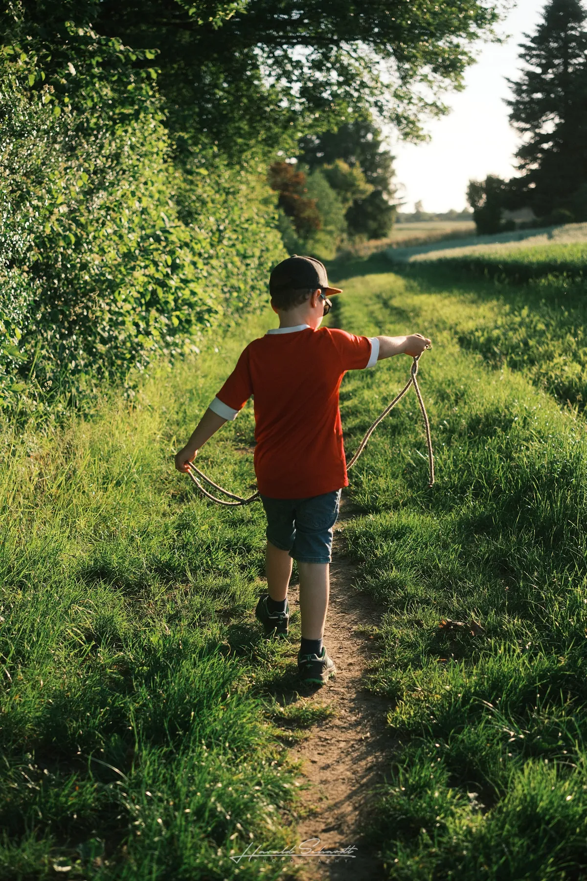 A young boy in a red shirt walks along a narrow path through green fields, holding a rope in the golden evening light.