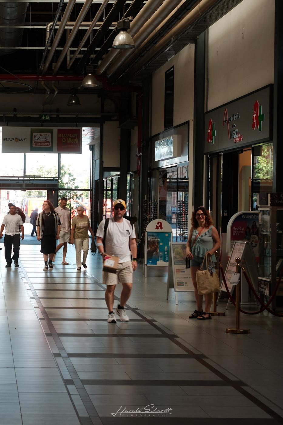 People passing through a covered shopping area with morning light filtering in from outside.