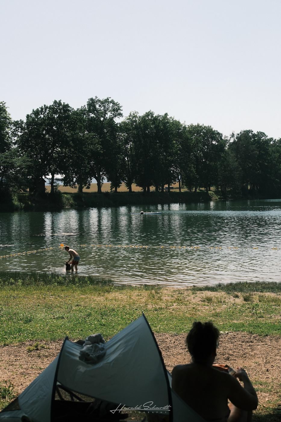 View across a calm lake with people relaxing on the shore beneath a line of trees.