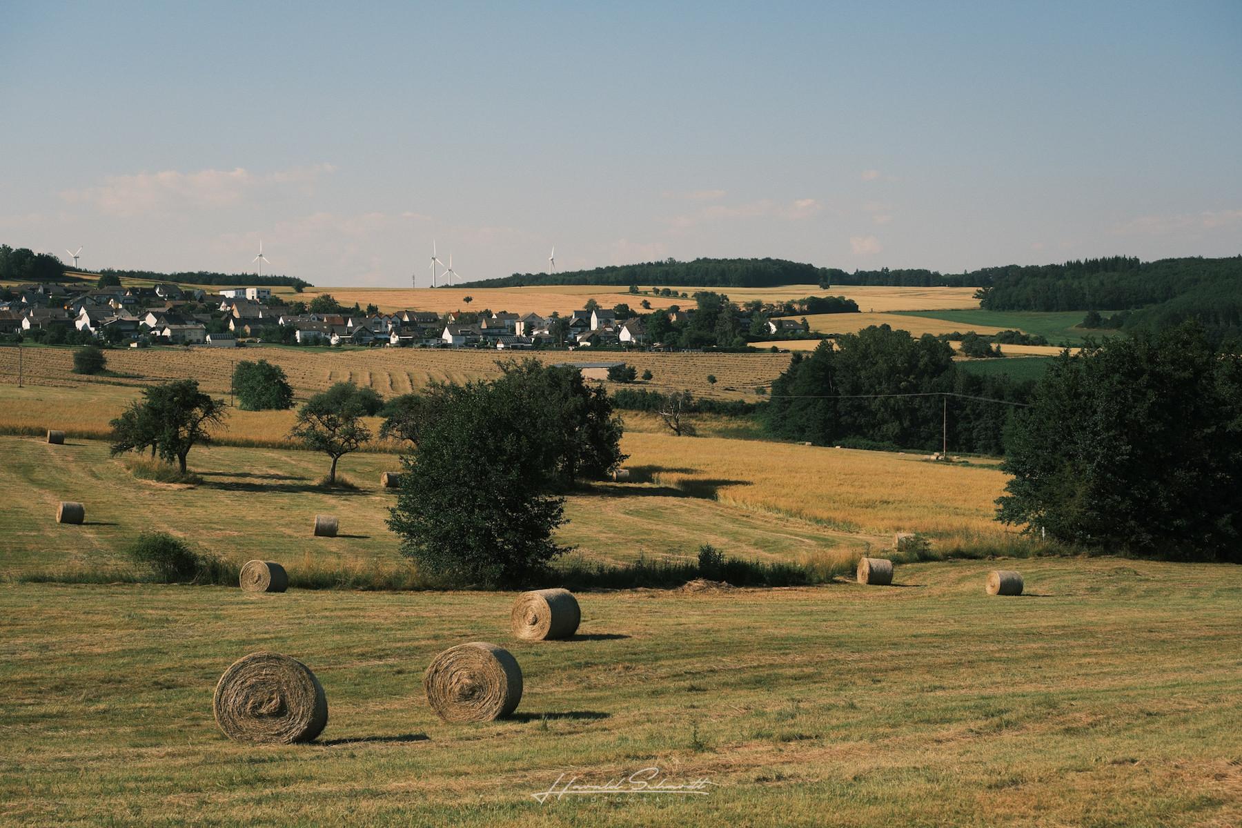 Wide view across rolling farmland with hay bales and a village on the horizon.