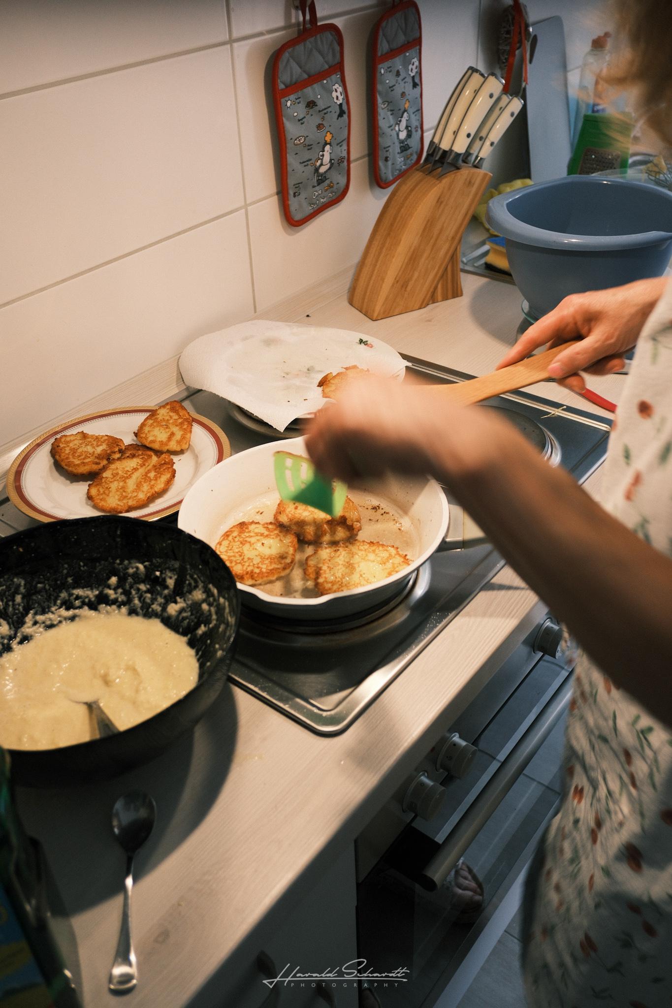 Potato pancakes frying in a pan on the stove, with a plate of finished ones and a bowl of batter alongside.