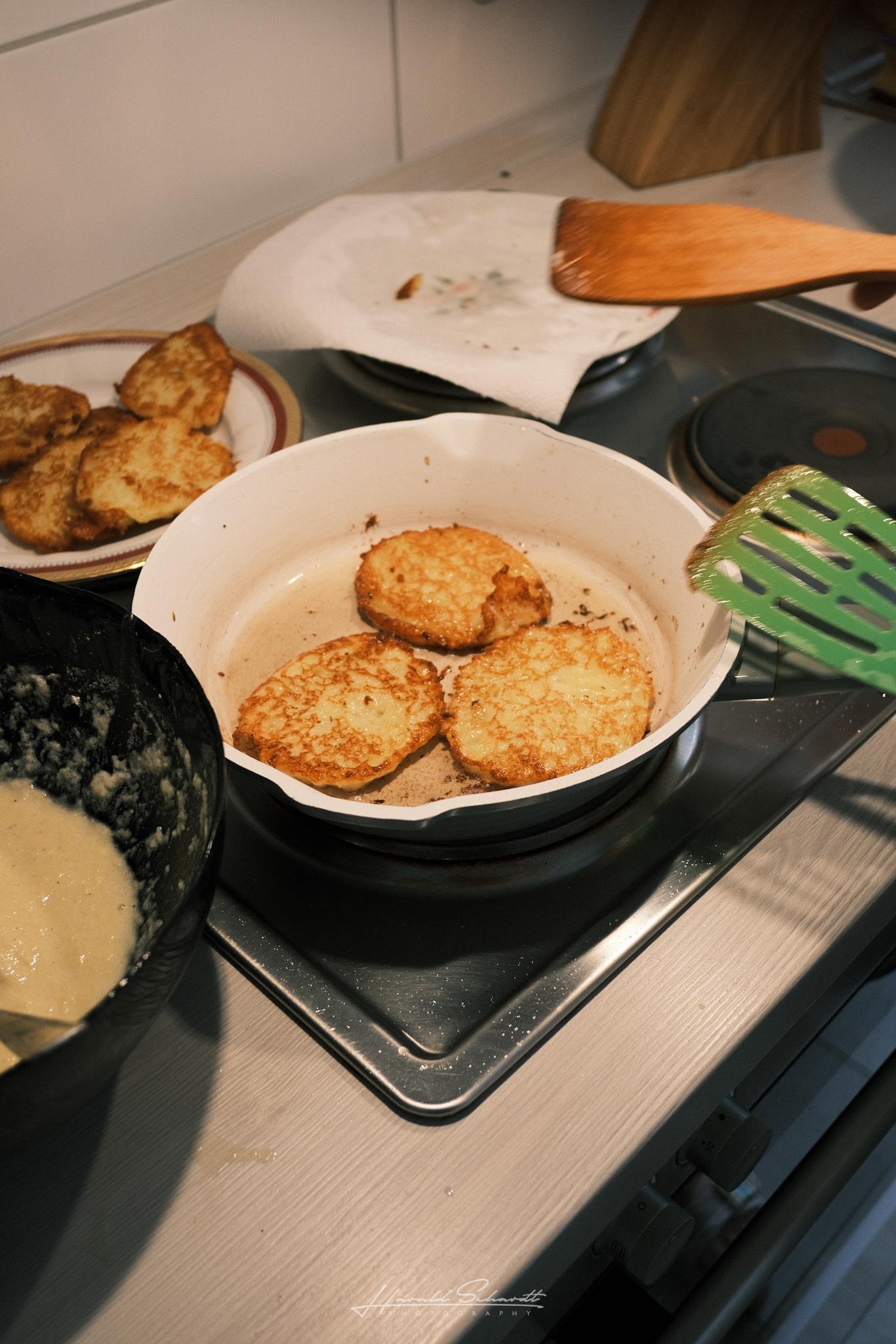 Three potato pancakes frying in a pan, with more finished ones on a plate nearby.