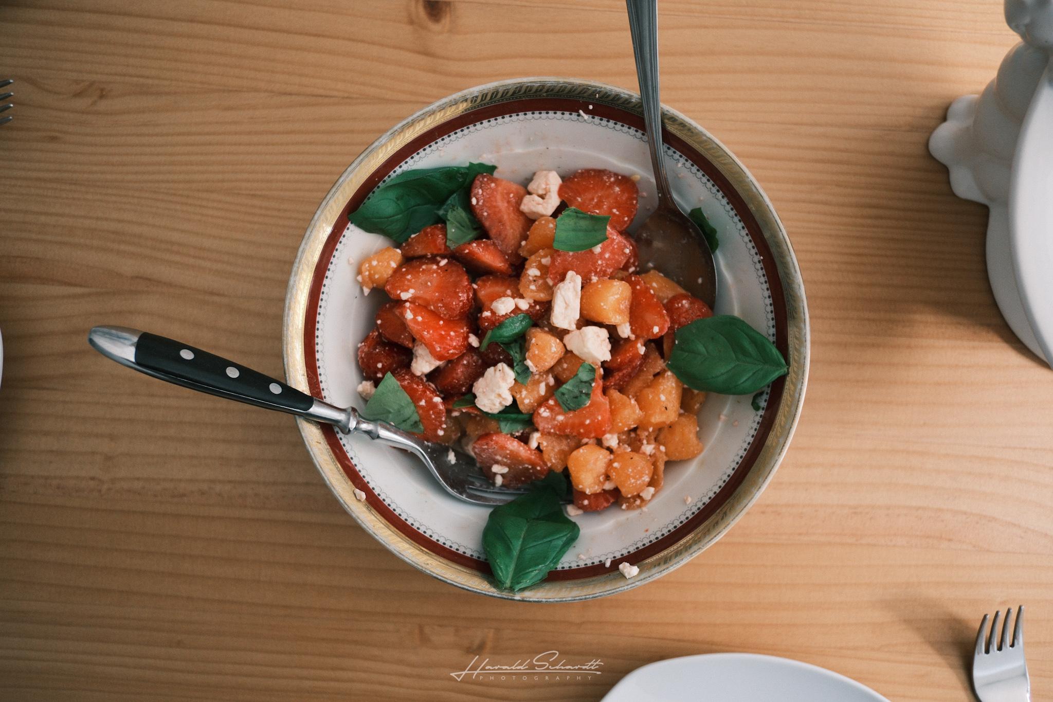 Overhead view of a bowl with diced strawberries, melon, feta, and basil leaves.