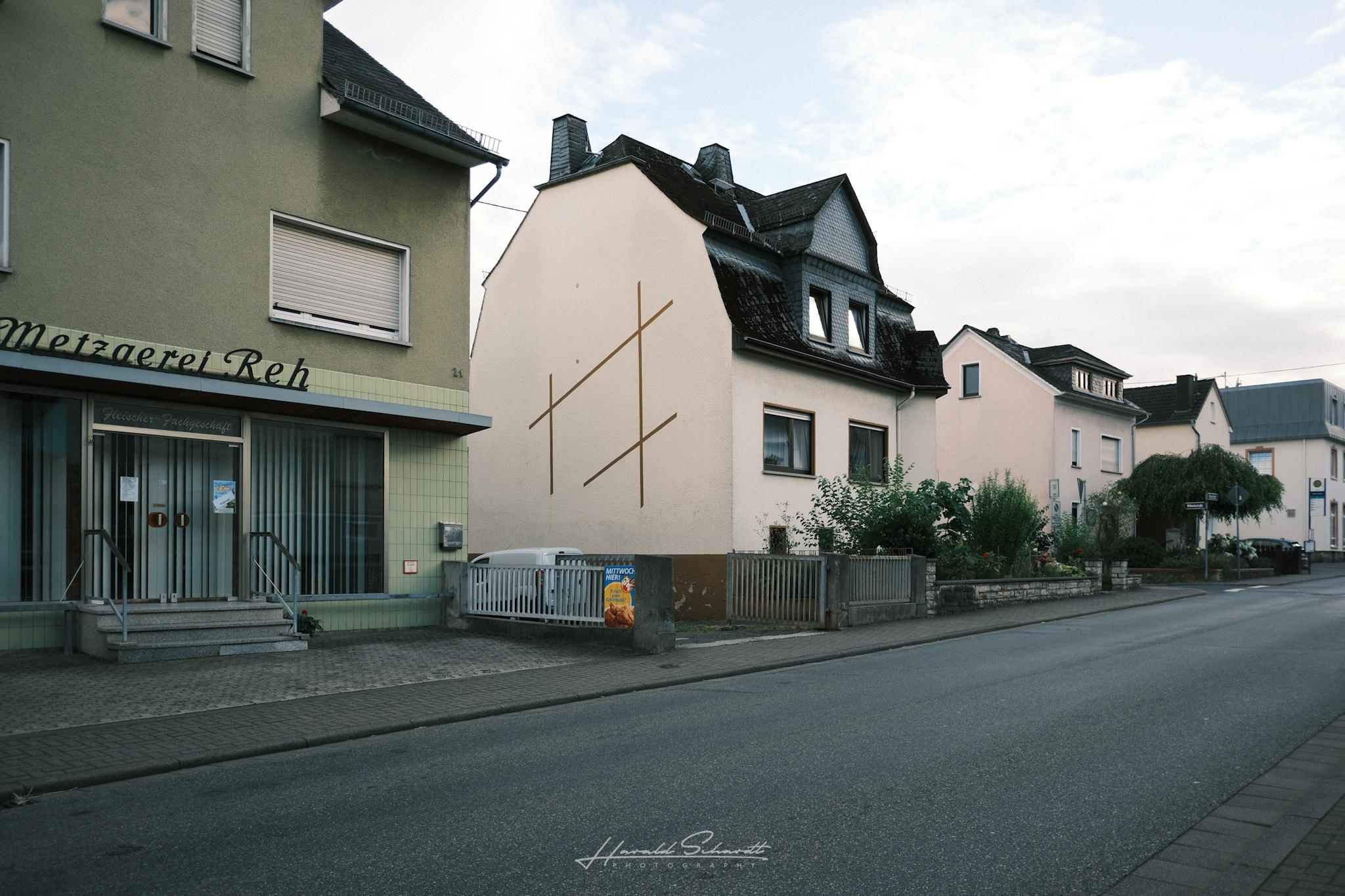 A quiet village street with older houses. One white wall is marked by a simple geometric line pattern.